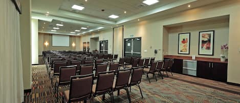 Un salón de conferencias con filas de sillas, una pantalla de proyector y una mesa de recepción con obras de arte.