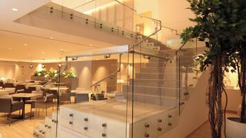 A modern hotel lobby with a glass staircase, seating area, and indoor plants.