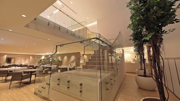 A modern hotel lobby with a glass staircase, dining area, and indoor plants.