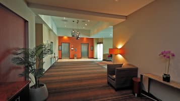 A hotel lobby with a carpeted floor, a reception desk, a potted plant, a sofa, and a staircase leading to the elevators.