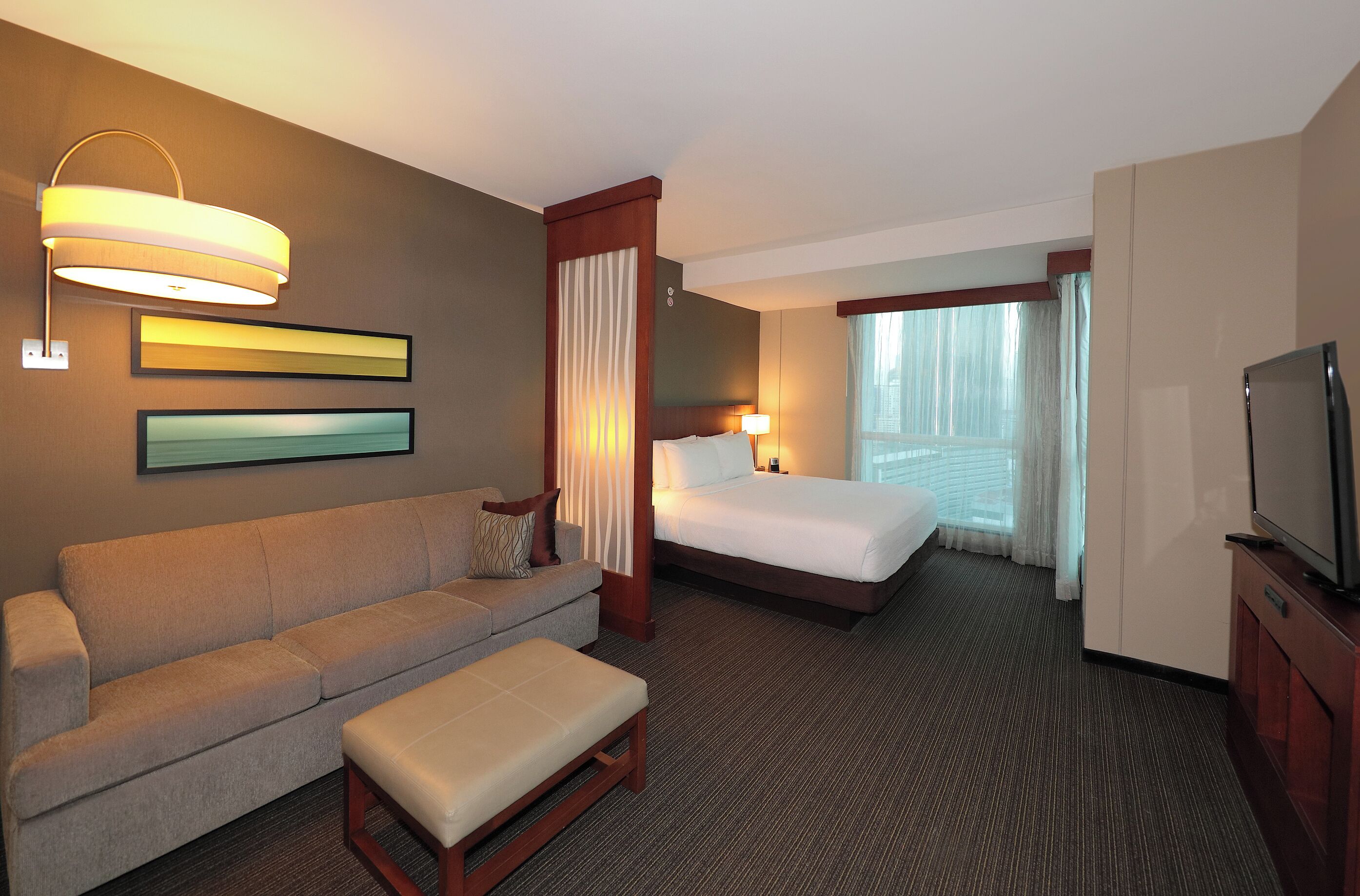 Una habitación de hotel con sofá, cama, televisión y ventana con cortinas.