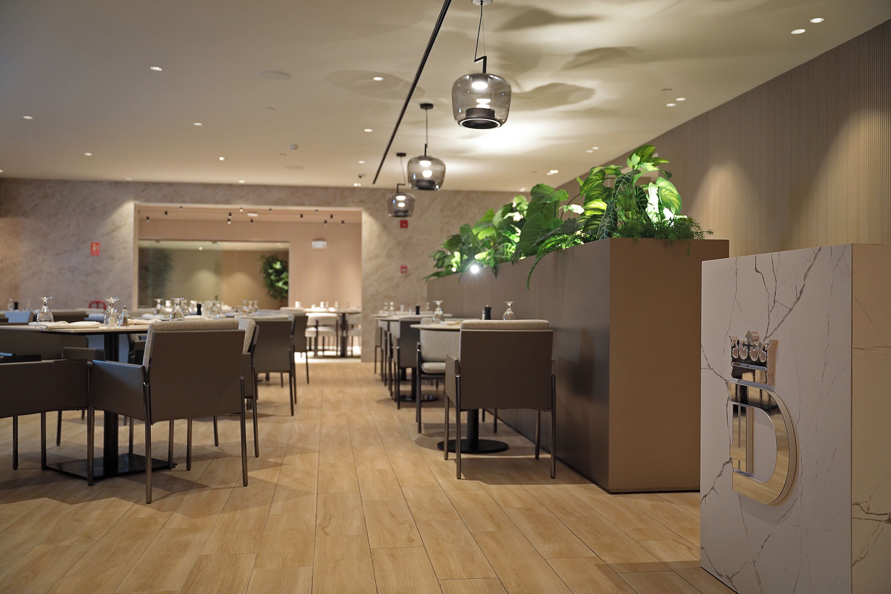 Un espace de restauration moderne doté d’un plancher en bois, de tables, de chaises, de luminaires suspendus et d’un mur où sont disposées des plantes.