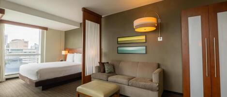 Habitación de hotel con cama, sofá, reposapiés, lámpara de pared y armario.