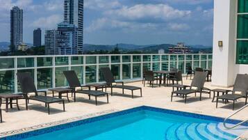 A rooftop pool with lounge chairs, a clear blue sky, and city buildings in the background.