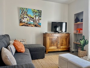 A living room with a gray sofa, a television on a wooden cabinet, a painting of trees, and a cactus plant.