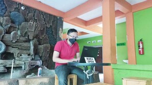 Un homme est assis à une table en bois, devant un ordinateur portable, dans une pièce dont un mur est en pierre et un autre est peint en vert, et sur lequel on peut lire l’inscription « WASTA FEL ».