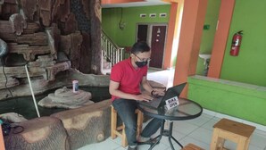 Un homme est assis à une table, devant un ordinateur portable, avec un tabouret en bois et une petite chaise en bois.
