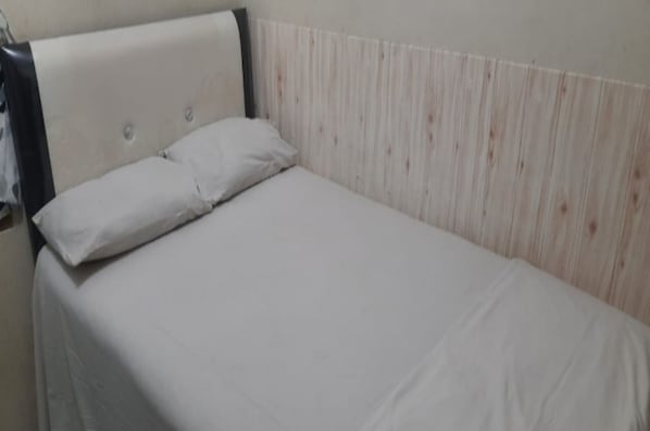 A single bed with a white mattress and pillows, a wooden headboard, and a plain wall.