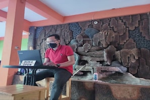 A person sitting at a table with a laptop labeled "BUKA OPEN" in a room with orange walls and a stone wall decoration.