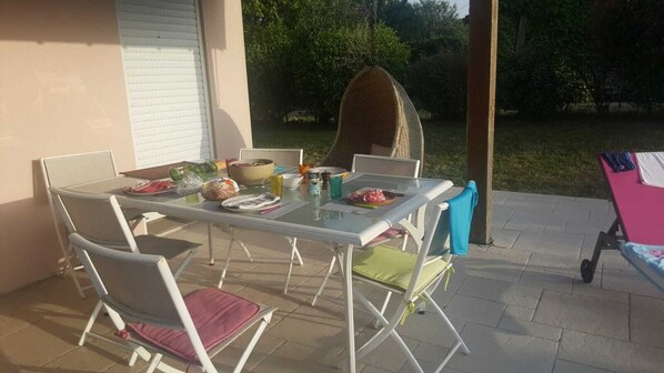 An outdoor patio with a glass table set for dining, surrounded by chairs with cushions.