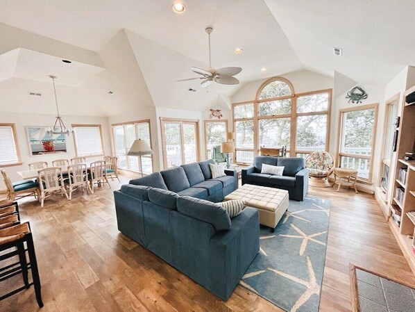 A spacious living room with a large sectional sofa, a smaller sofa, and a coffee table. There is a dining area with a table and chairs, and a bookshelf in the corner.