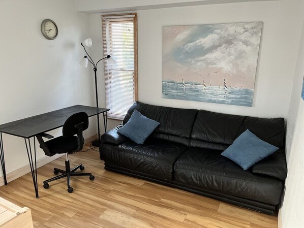A room with a black leather sofa, a desk with a chair, a floor lamp, a painting of birds on water, and a clock on the wall.