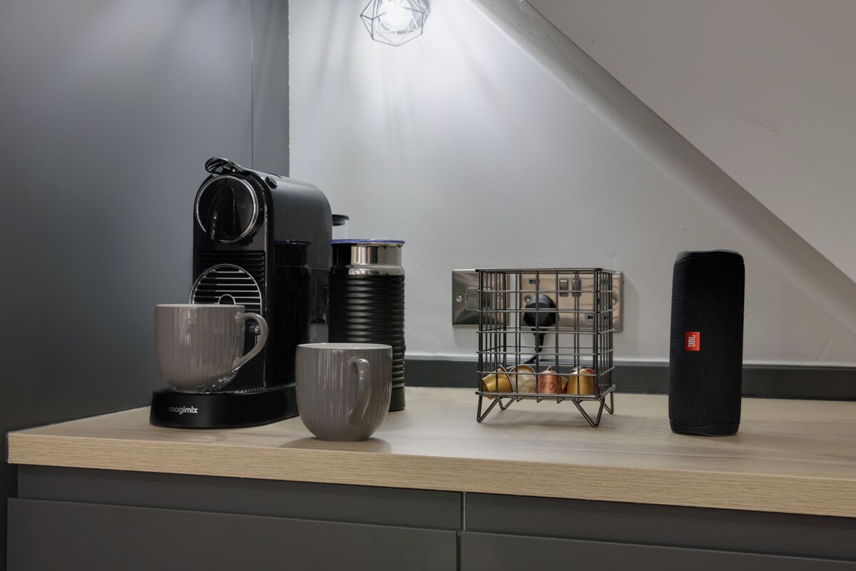 Un comptoir de cuisine avec une machine à café, deux tasses, une bouilloire, un petit présentoir métallique pour tasses et un haut-parleur noir.