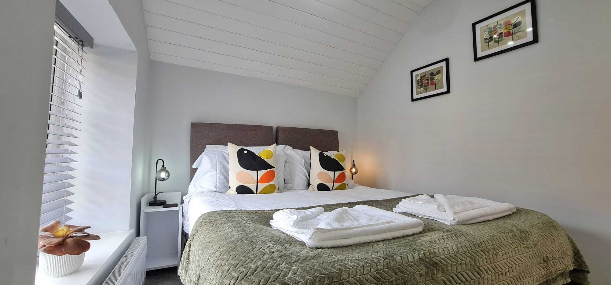 A neatly made bed with white linens and colorful pillows, a bedside lamp, and a folded towel on the floor.