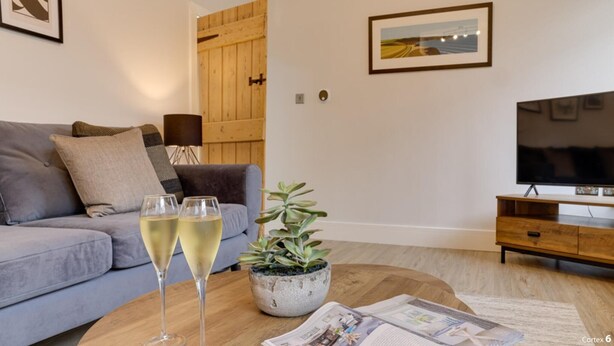 A living room with a grey sofa, a wooden coffee table with two glasses of white wine, a potted plant, and a television on a wooden stand. - Beam End Snainton - Modern 2-bedroom Stone Cottage. Pet Friendly. (Snainton)