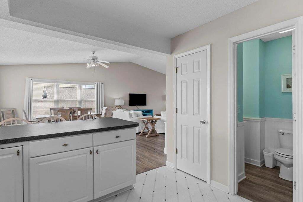 A kitchen with white cabinets, a black countertop, and a ceiling fan. There is a dining area with a table and chairs, and a bathroom with a toilet and sink.