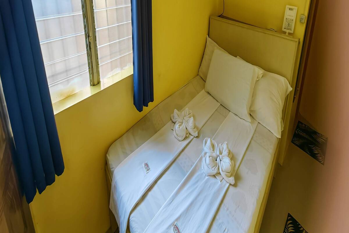 Habitació petita amb un llit individual, una paret groga, una finestra amb cortines blaves i un cartell de WiFi penjat a la paret.