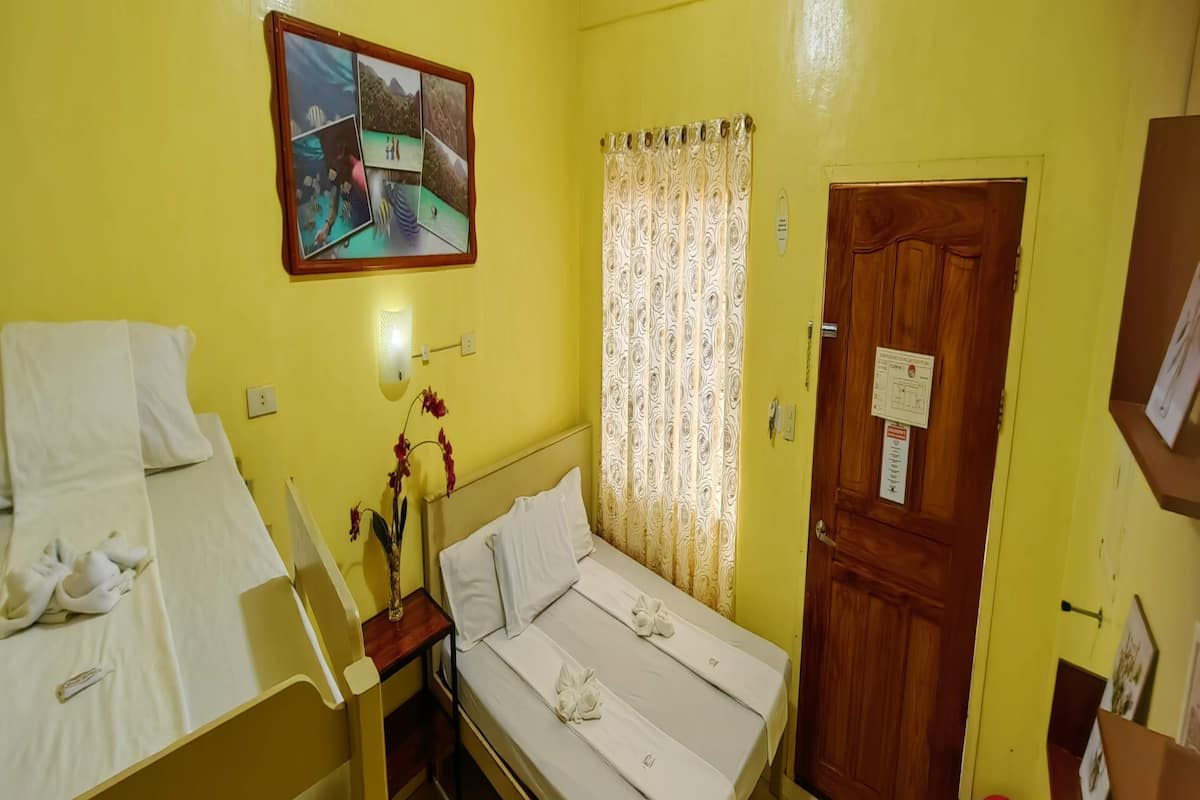 Habitació amb dos llits, un panell de capçalera de fusta, una taula petita amb un ram de flors, una finestra amb cortines estampades i una porta amb un cartell.