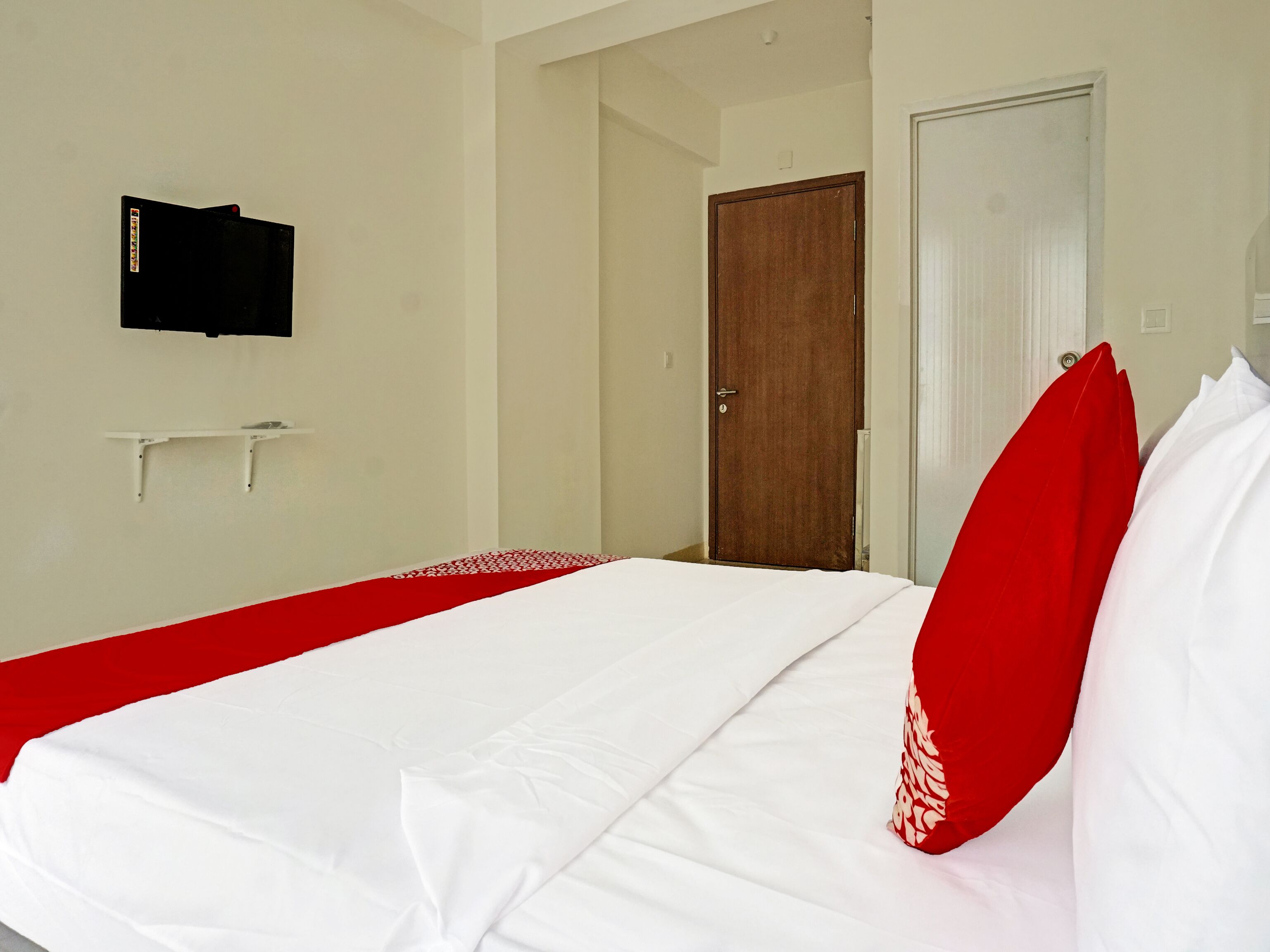 A hotel room with a bed, a television mounted on the wall, a wooden door, and a white door.