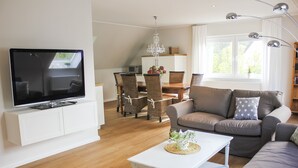 A modern living room with a flat-screen TV, a white coffee table, a brown sofa, wicker chairs, and a dining area with a chandelier. - Deichkind * Holiday apartment on the North Sea in Husum * At home by the sea (Husum)