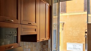 A kitchen with wooden cabinets, a gas stove, a sink, and a window with a view of a building labeled "VIA DELLE COLONNETTE". - Stay in the heart of Rome (Roma)