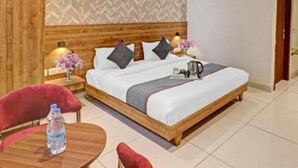 A hotel room with a wooden headboard, a bed with pillows, a bedside table with a lamp, a small table with a water bottle, and a ceiling fan. - Hotel Golden Glory (New Delhi)