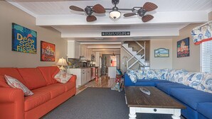 A living room with a red sofa, blue sofa, and a wooden coffee table. There is a ceiling fan, kitchen area, and a staircase leading to another level. - Beautiful townhouse with outdoor pool  Free WiFi, linens included and right at the beach. (Ocean City)