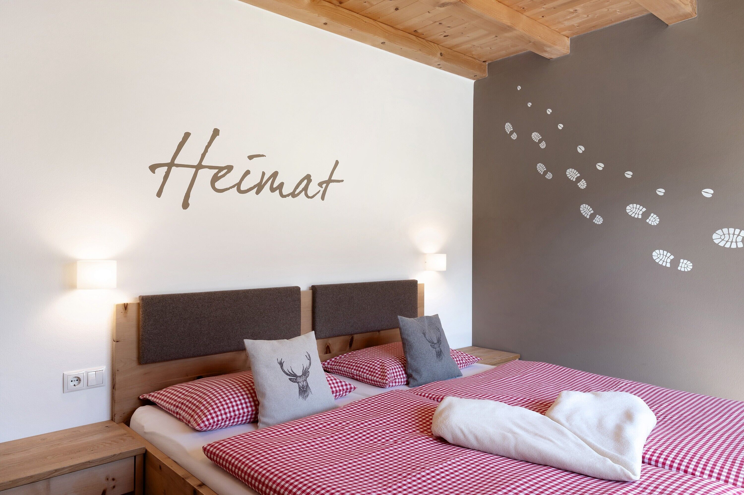 Un lit avec une literie à carreaux, une tête de lit ornée d’un coussin à motif de cerf et un mur sur lequel est écrit le mot « Heimat » ainsi qu’un autocollant mural.