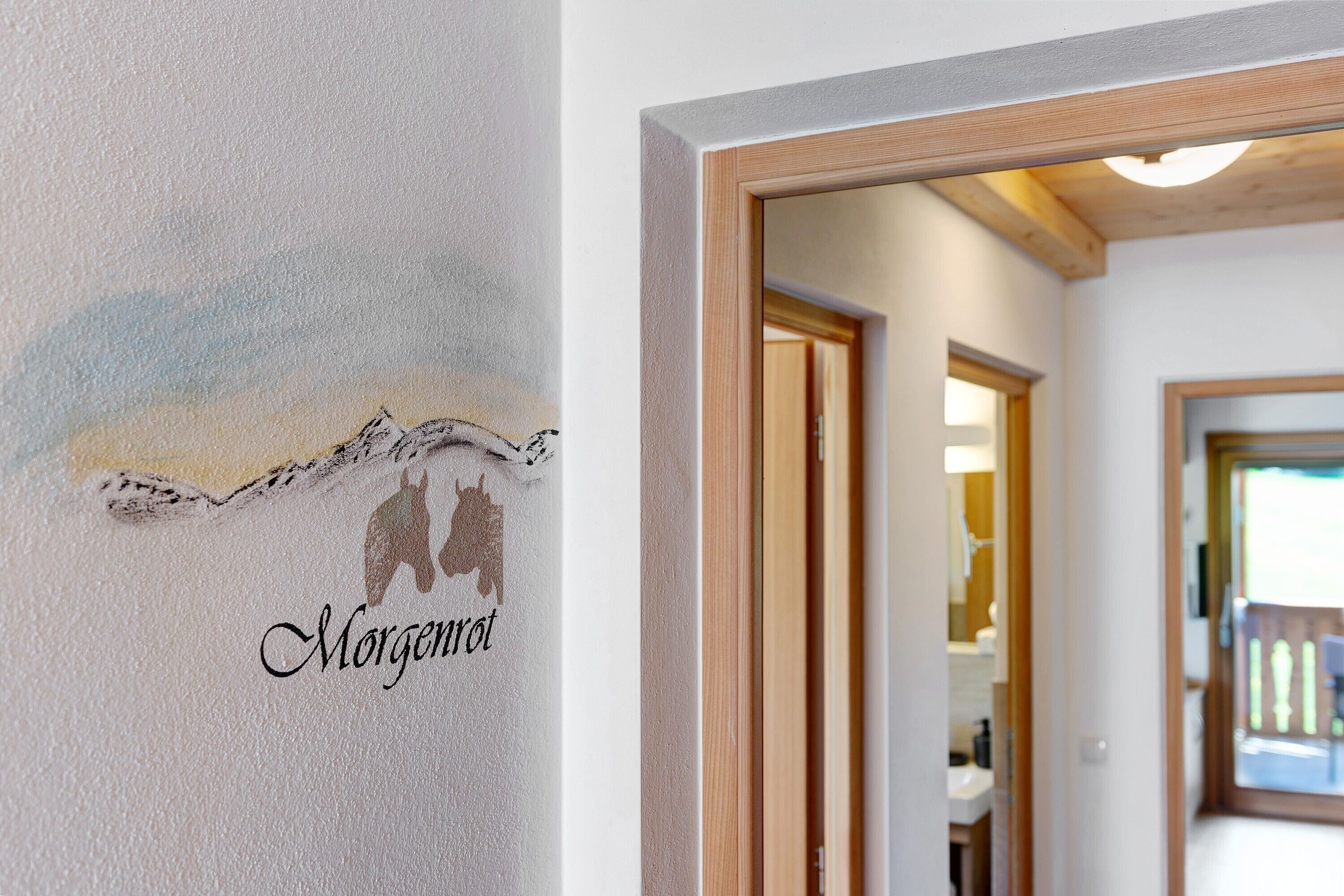 Um corredor com moldura de porta de madeira, uma placa na parede com silhueta de montanha e a palavra 'Morgenrot', e uma visão de um quarto com janela.