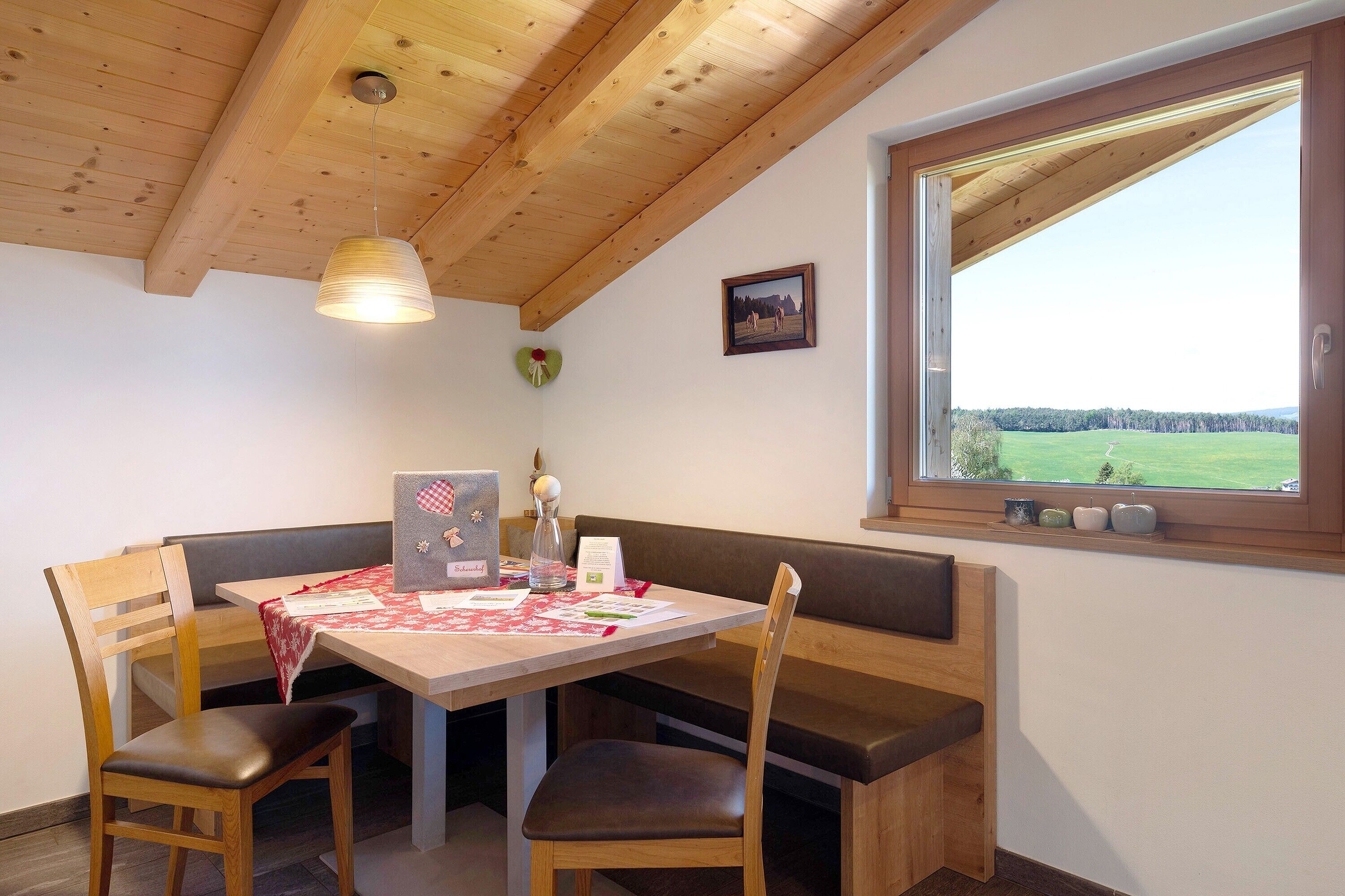 A dining area with a wooden table and bench, a hanging light fixture, a framed picture on the wall, and a window with a view of a green landscape.