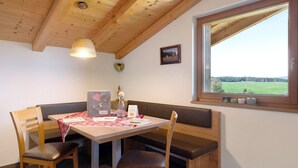 Ein Essbereich mit Holztisch und Bank, einer Hängelampe, einem gerahmten Bild an der Wand und einem Fenster mit Blick auf eine grüne Landschaft.
