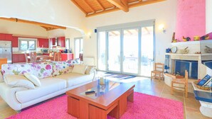 A spacious living area with a white sofa, a wooden coffee table, and a pink rug. There is a kitchen in the background with red cabinets and a dining area with chairs. - Villa Kalamia in Kalami (Corfu)