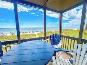 Un espace extérieur abrité, avec une table et des chaises, donnant sur une plage parsemée de parasols et fréquentée par des gens.