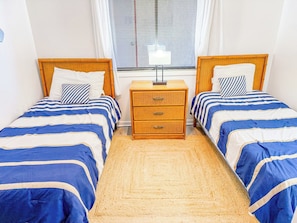 A bedroom with two single beds, a nightstand, a lamp, and a window with blinds. - Tilghman Beach and Racquet Club 209 (North Myrtle Beach)