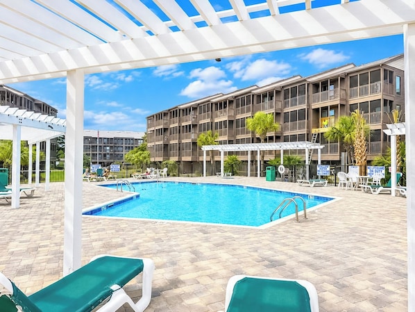 A swimming pool area with lounge chairs, a covered seating area, and a multi-story building in the background. - Tilghman Beach and Racquet Club 209 (North Myrtle Beach)