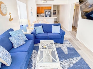 A living room with a blue sofa set, a coffee table, and a kitchen area in the background.