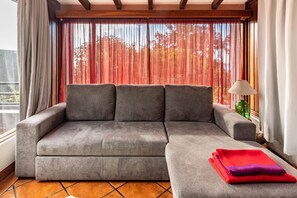 A gray sofa with a matching ottoman, a green glass bottle, and a lamp on a wooden floor with a view of trees through red curtains.