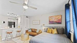 A modern bedroom with a large bed, a sofa, and a small kitchen area with a refrigerator and bar stools. - Stylish Modern Studio Close to Downtown and Domain (Austin)