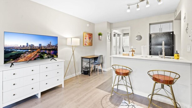 A modern kitchen with a large TV, white cabinets, and a bar area with stools. - Stylish Modern Studio Close to Downtown and Domain (Austin)