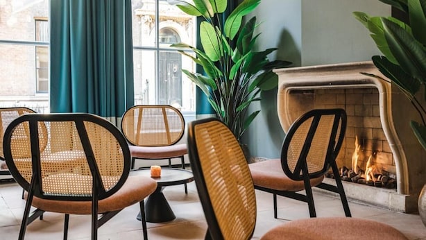 A cozy seating area with wicker chairs and a large plant, a fireplace, and a view of a building through a window with blue curtains. - Hotel Halbert (Groningen)