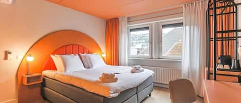 A hotel room with a bed, a desk, a chair, a window with curtains, and a radiator.