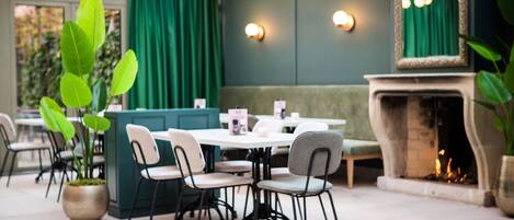 A dining area with a green booth, white tables, and chairs. There is a fireplace and a potted plant.