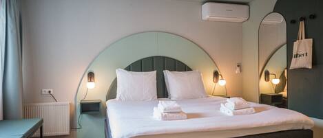 A neatly made bed with white linens and towels, a bedside table, a wall-mounted air conditioner, and a mirror with a towel hanging on it.