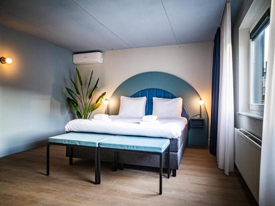 Une chambre d’hôtel avec un lit, une table de chevet, une lampe, une plante en pot et une fenêtre avec des rideaux.