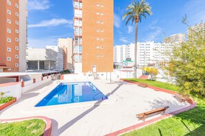 Un área de piscina con una piscina azul cristalina, rodeada por una terraza blanca, con una palmera y edificios al fondo.