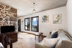 Una sala de estar con pared de piedra, piso de madera, televisor de pantalla plana, un sofá y dos ventanas.