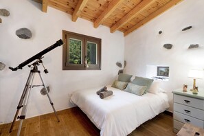 Un dormitorio con un telescopio, una cama con almohadas, un aparador y una ventana con vistas a árboles.