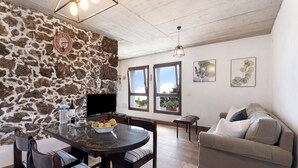 Un coin repas avec un mur en pierre, une table ronde en verre et des chaises. On aperçoit un canapé et un téléviseur au fond.