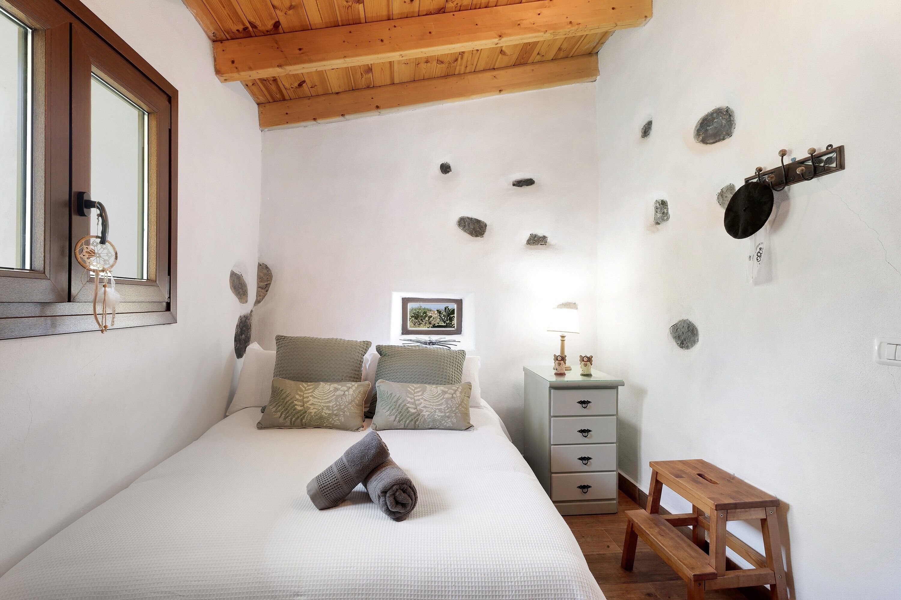 A bedroom with a bed, pillows, a bedside table with a lamp, a wooden bench, and a wall with rocks.