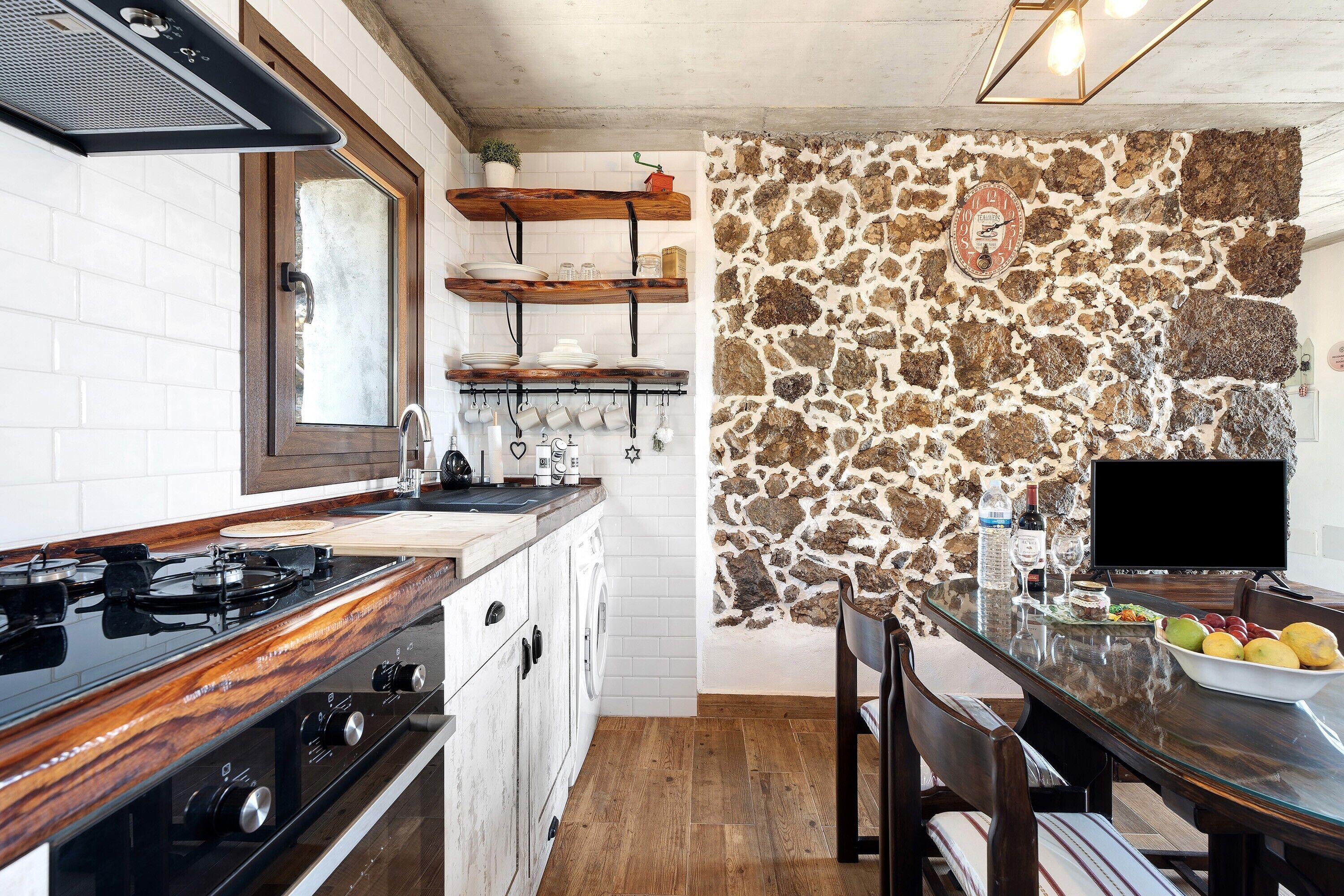 Une cuisine avec un mur en pierre, des étagères en bois, une cuisinière et un coin repas comprenant une table et des chaises.